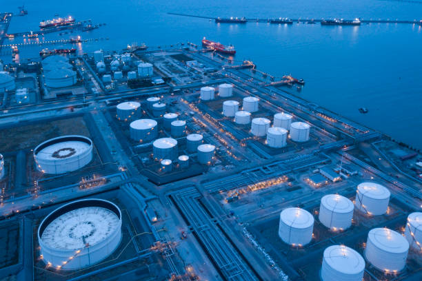 An aerial view of a bustling oil refinery and tank farm, showcasing a complex network of pipelines, storage tanks, and processing facilities. The image captures the industrial scale of the operation, highlighting the importance of petroleum in the global economy. The vibrant blue water surrounding the facility adds a striking contrast to the grey metal structures, creating a compelling visual. This photo is ideal for illustrating the infrastructure of the oil and gas industry, energy production, and industrial landscapes.